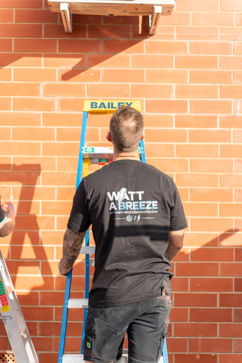Watt A Breeze technician installing an outdoor air conditioning unit on a ladder